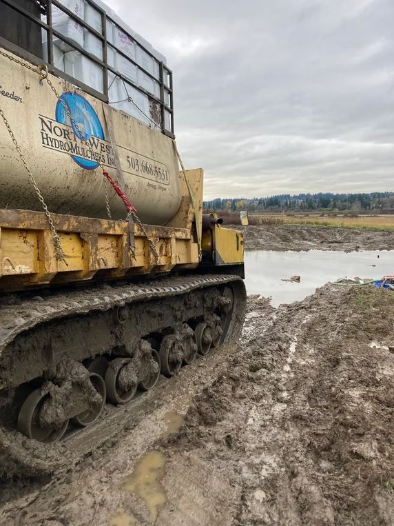 Yellow tracked construction vehicle parked in thick mud near a body of water under a cloudy sky.