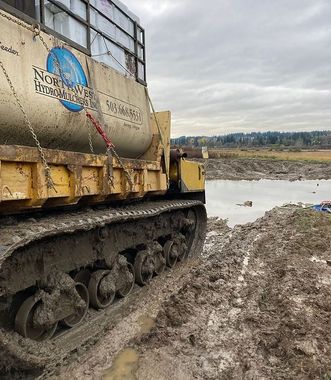Yellow tracked construction vehicle parked in mud next to a water-filled trench in an open, overcast field.