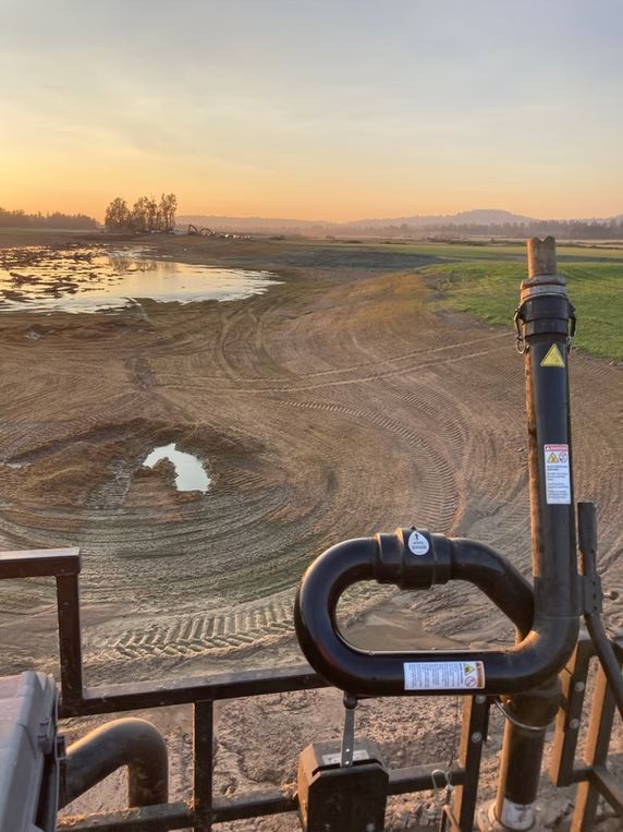 View from a tractor cab overlooking a muddy, partially flooded field at sunset, with machinery parts in the foreground.