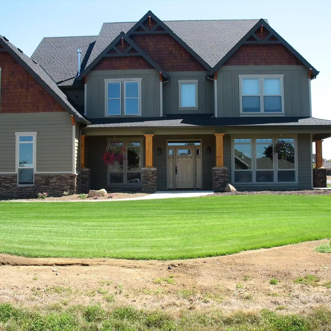 A two-story suburban house with brown wood shingles, green siding, stone accents, and a wide front porch with a green lawn.