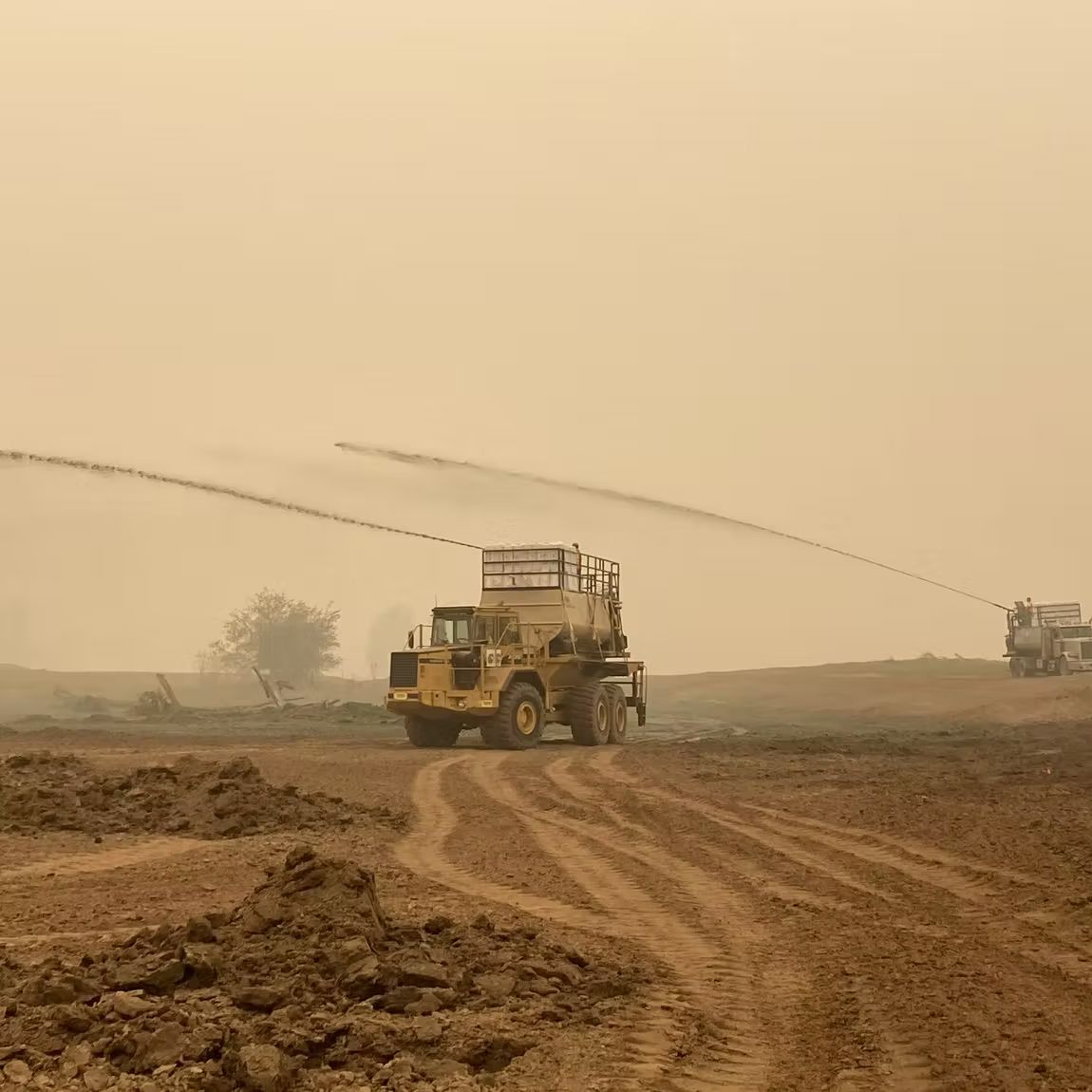 Yellow industrial water trucks spraying water onto dry, dusty earth on a hazy, overcast day.