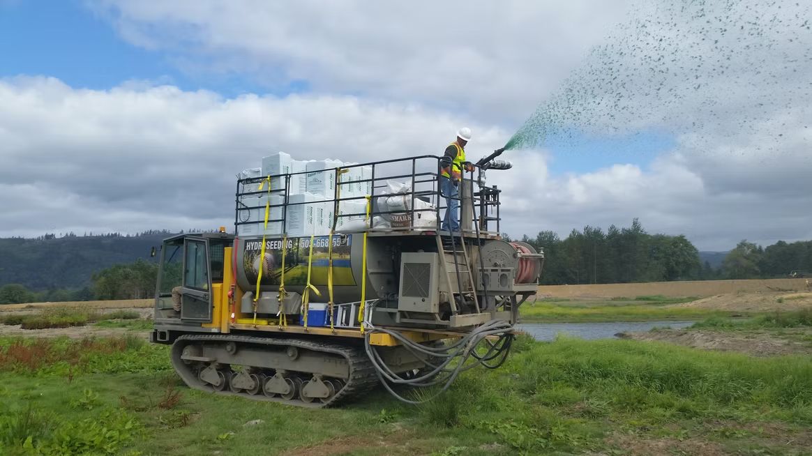 A worker on a tracked utility vehicle sprays a green mulch mixture across a field near a body of water.