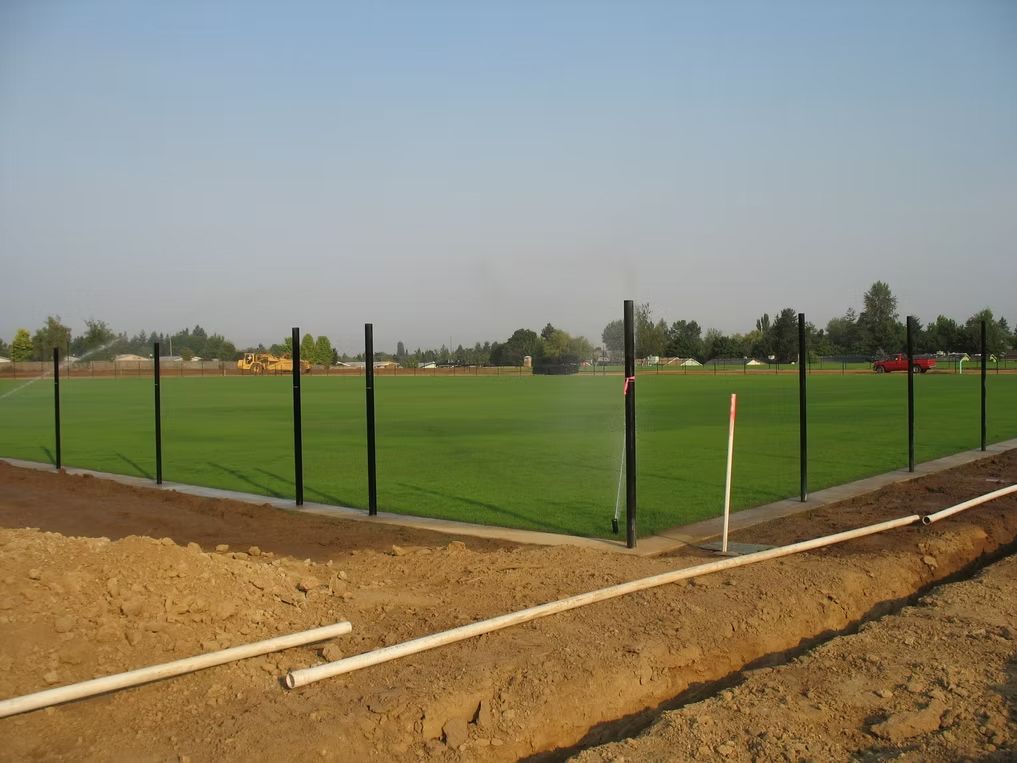 A rectangular sports field with bright green grass enclosed by a black fence, with construction piping in the foreground.