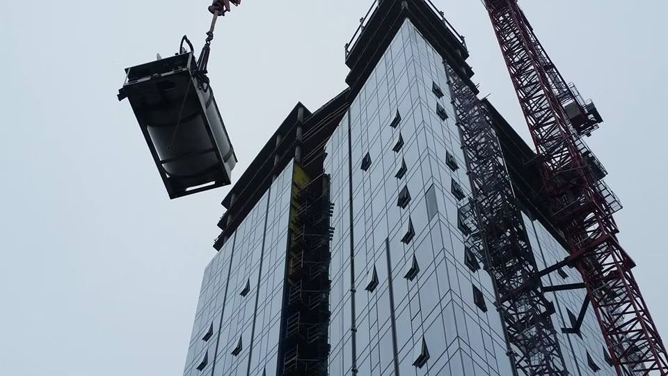 A large construction crane lifts a mechanical unit toward the top of a modern, glass-paneled skyscraper.