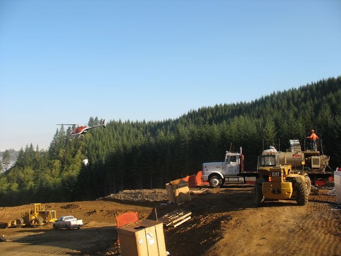 A helicopter flies over a construction site with heavy machinery, trucks, and a forested hillside in the background.