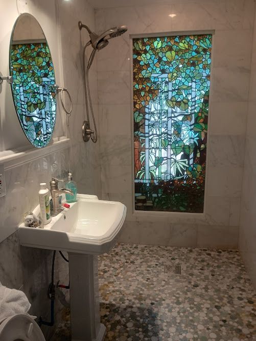 A bathroom with a sink , mirror , shower and stained glass window.