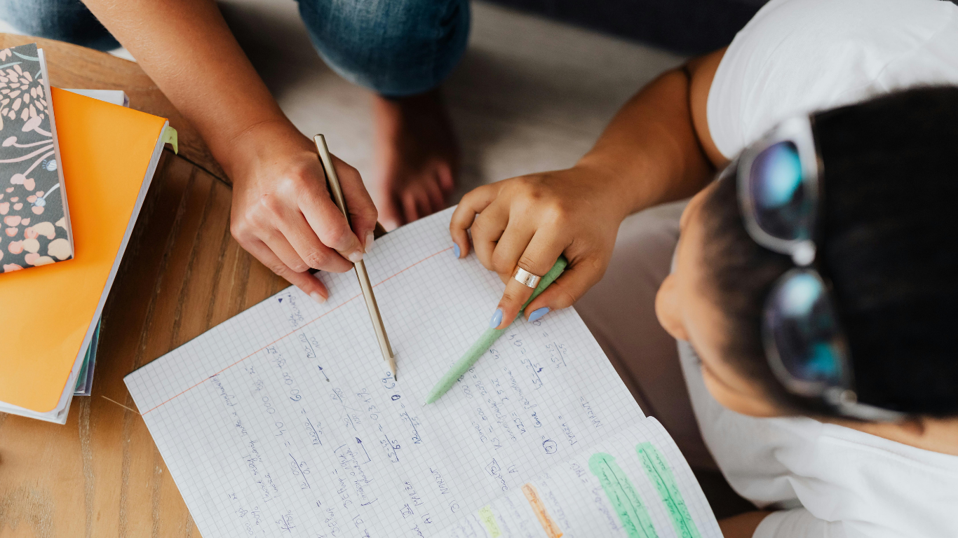 Two people studying a document with pens, one with glasses, books on a table.