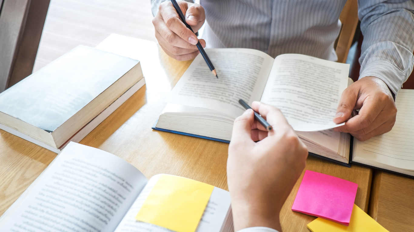 Hands pointing at open books on a desk, with sticky notes.