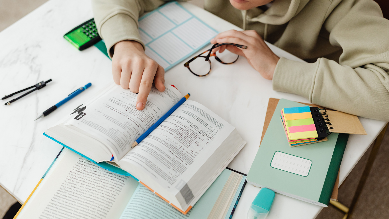 Person studying at a desk, pointing at an open book with glasses, pens, and notebooks.