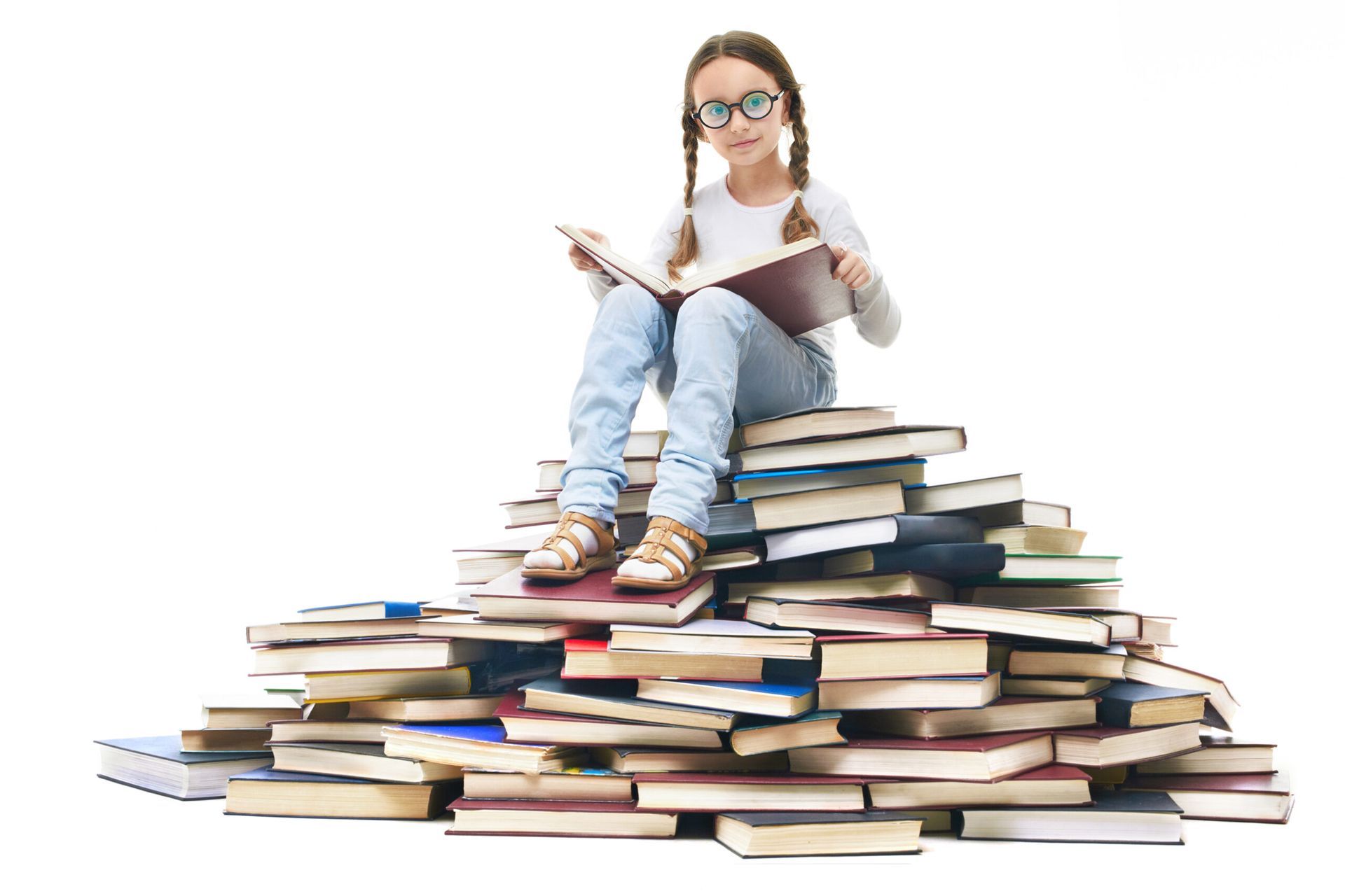 A young person with glasses and pigtails reads a book while sitting atop a large, disorganized mound of books.