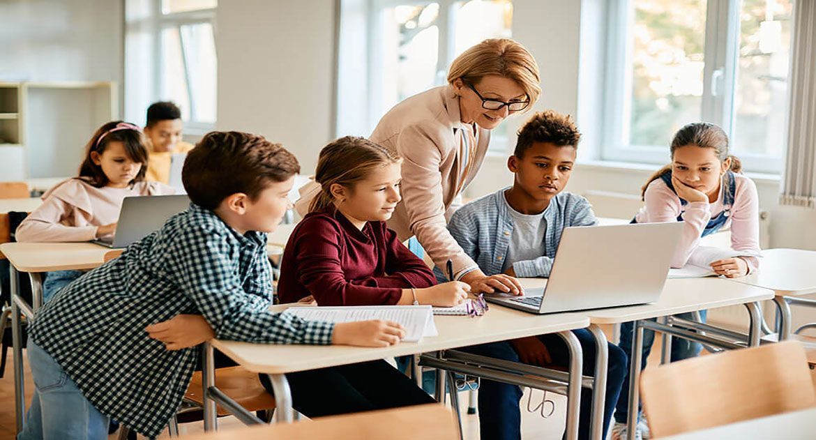 A teacher assists students with a laptop in a brightly lit classroom, with children sitting and working at desks.