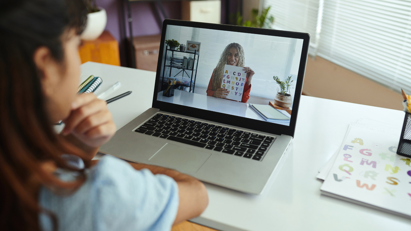 A student sits at a desk with a laptop, watching a virtual lesson with a teacher holding an alphabet learning card.