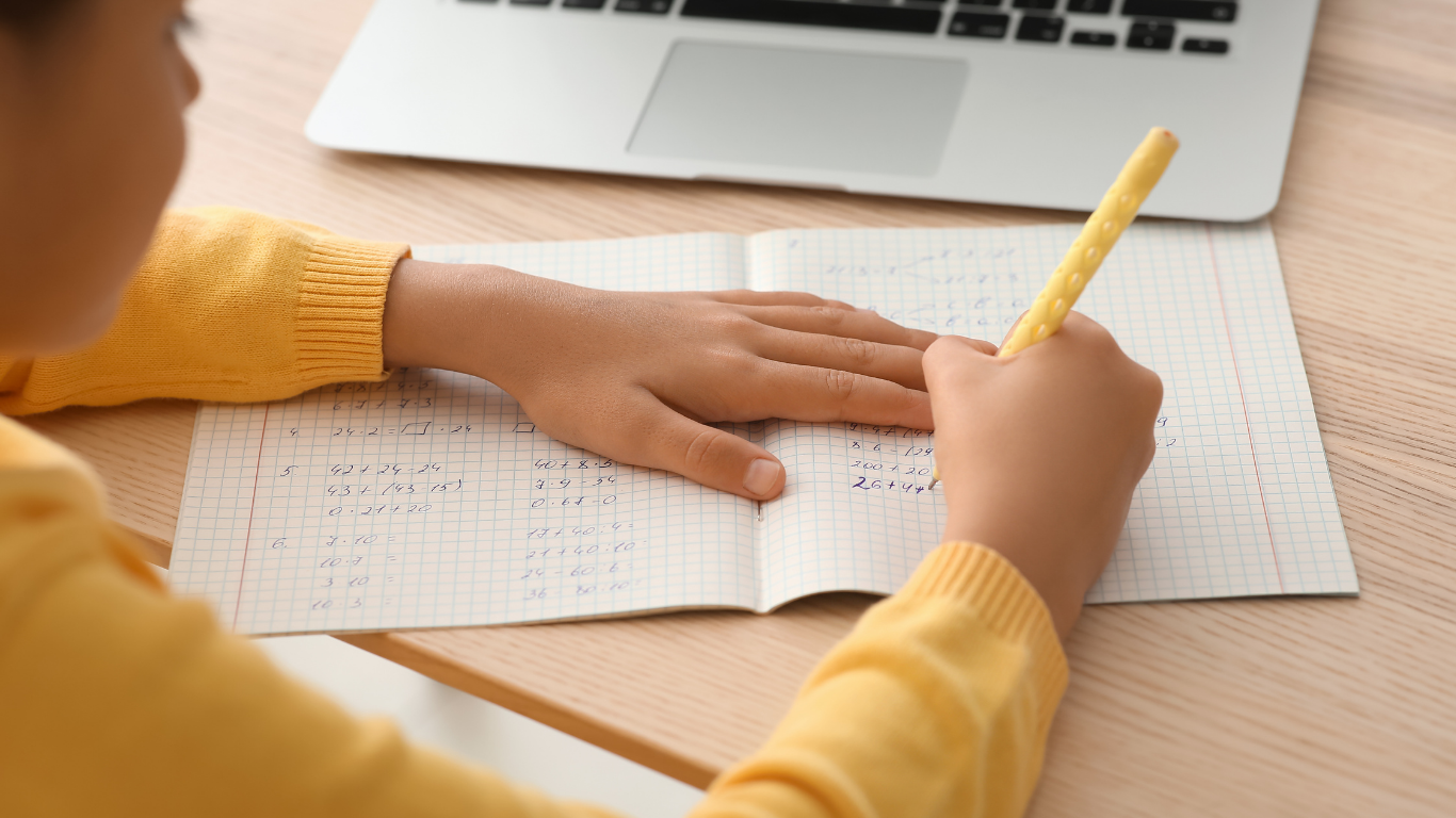 A person wearing a yellow sweater writes in a graph paper notebook at a desk with a laptop nearby.
