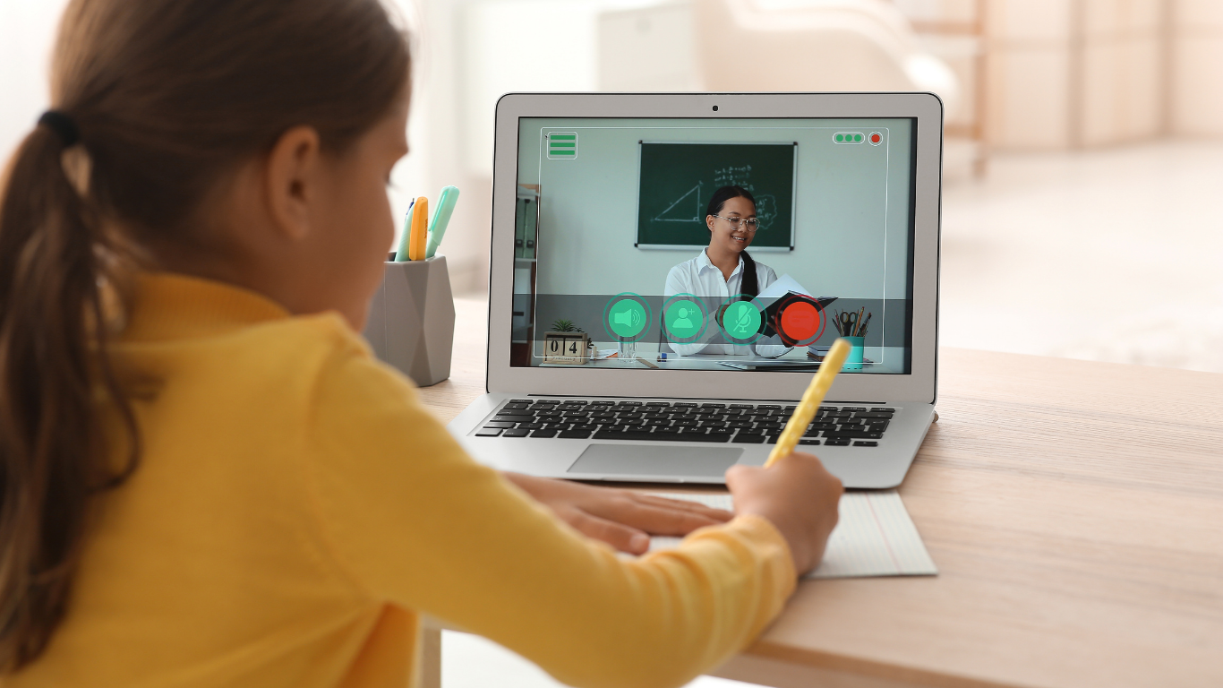 A student in a yellow shirt writes on paper while watching an online lesson on a laptop.