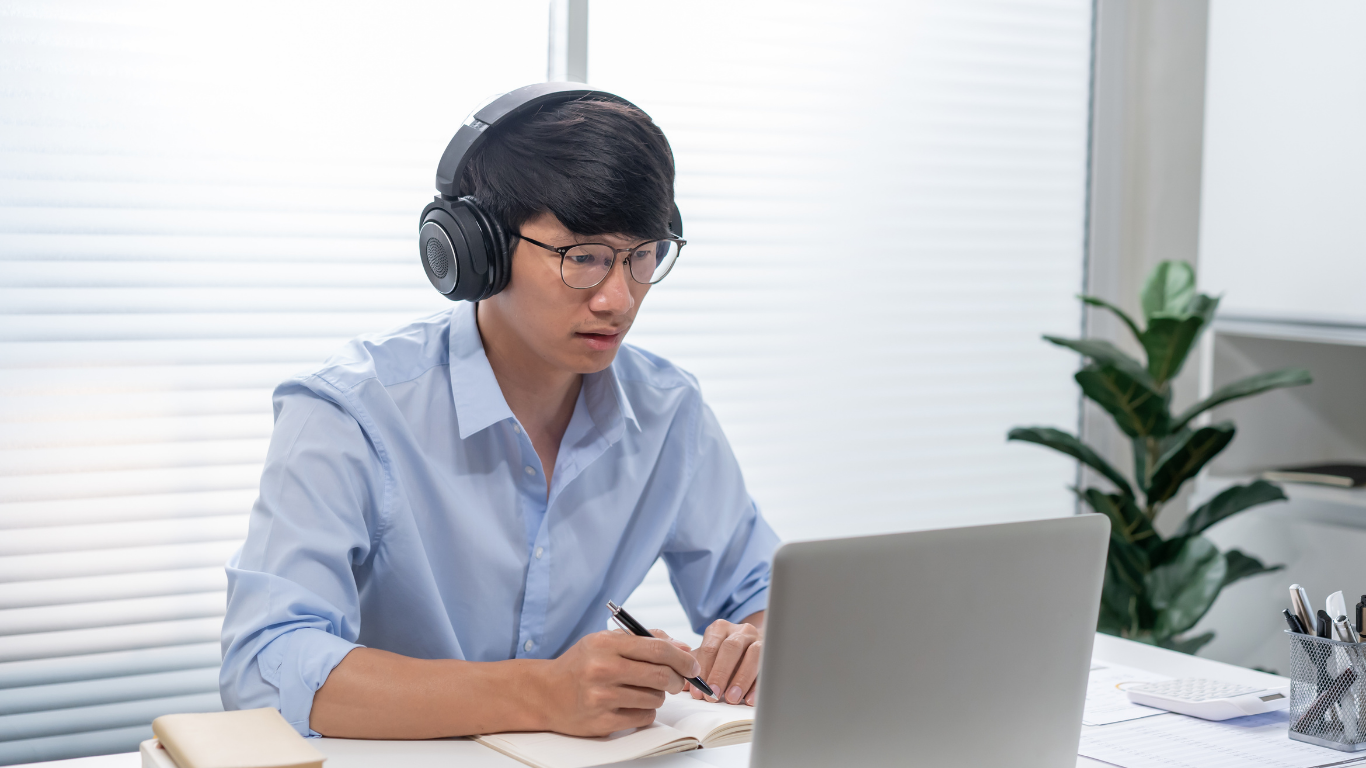 A person wearing headphones and glasses writes in a notebook while looking at a laptop in a bright office.