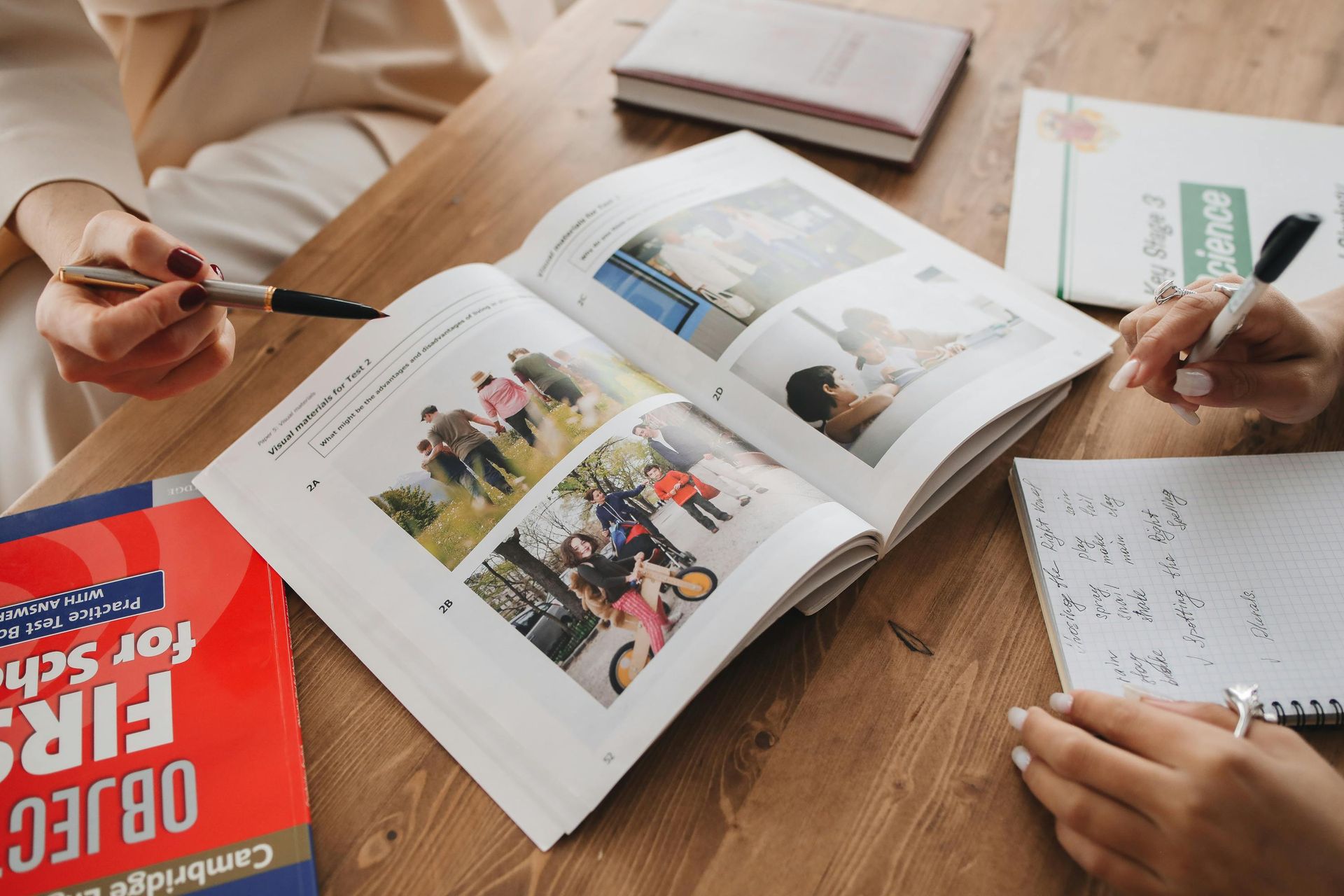 Two people study a textbook with photos, taking notes at a wooden table with books.