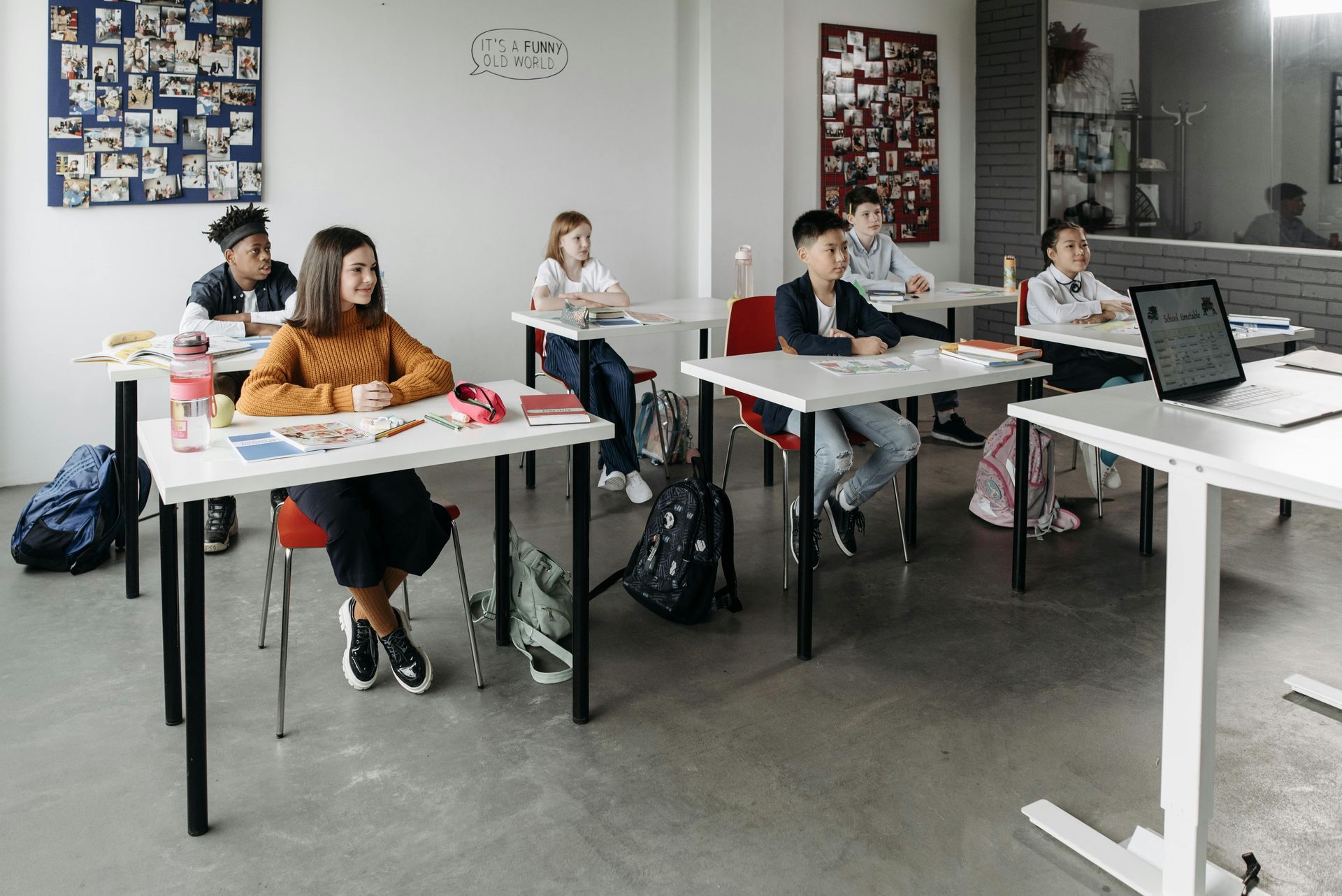 Children seated at desks in a classroom, some working, some looking forward. Brightly lit room.