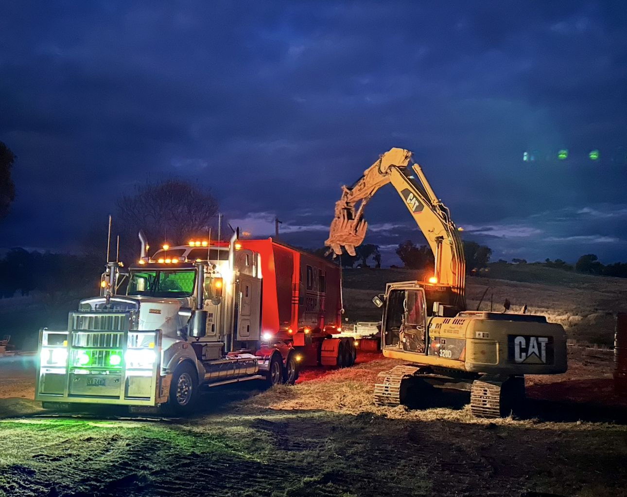 A yellow excavator loading scrap on a white truck with orange bins at night.