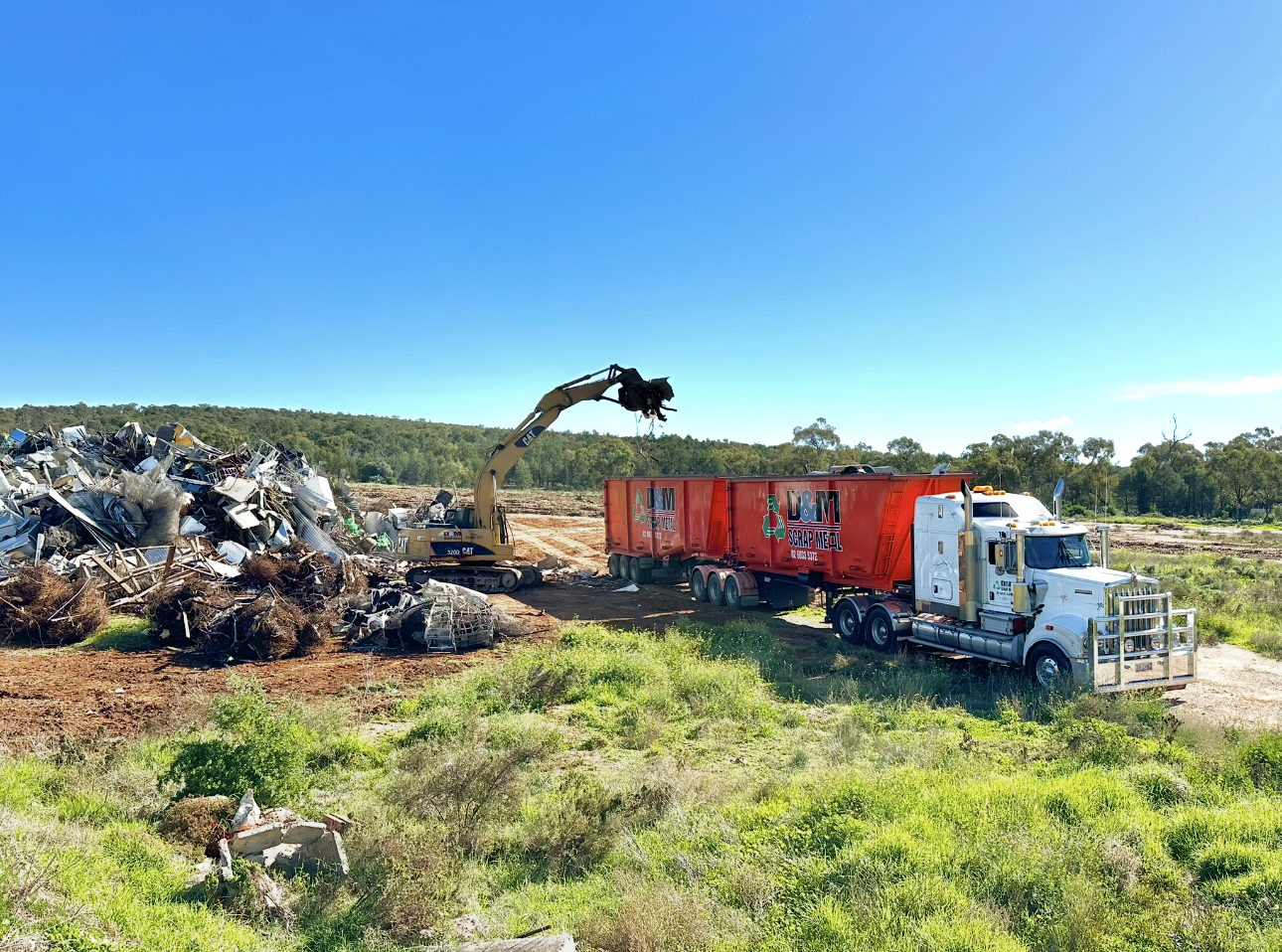 A yellow excavator with a claw loading scrap metal on orange bins on the back of a white truck.