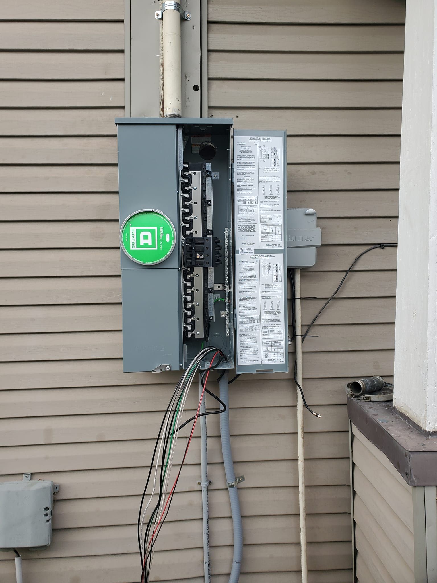 Exterior electrical panel on a beige-sided house, door open, with wires exposed.