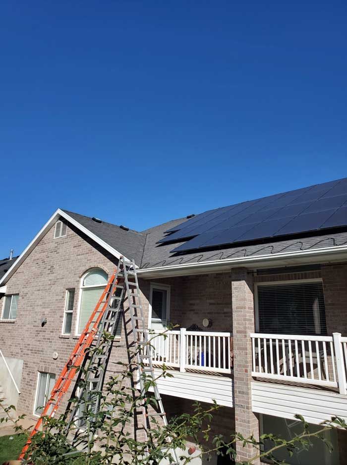 Brick house with installed solar panels on roof; orange ladder leans against the building. Bright blue sky.