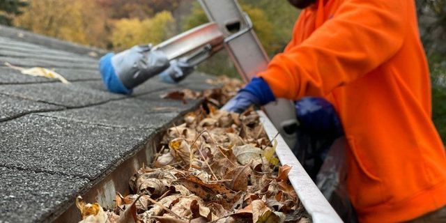 Worker in orange jacket clearing leaves from a roof gutter with a blue vacuum tool