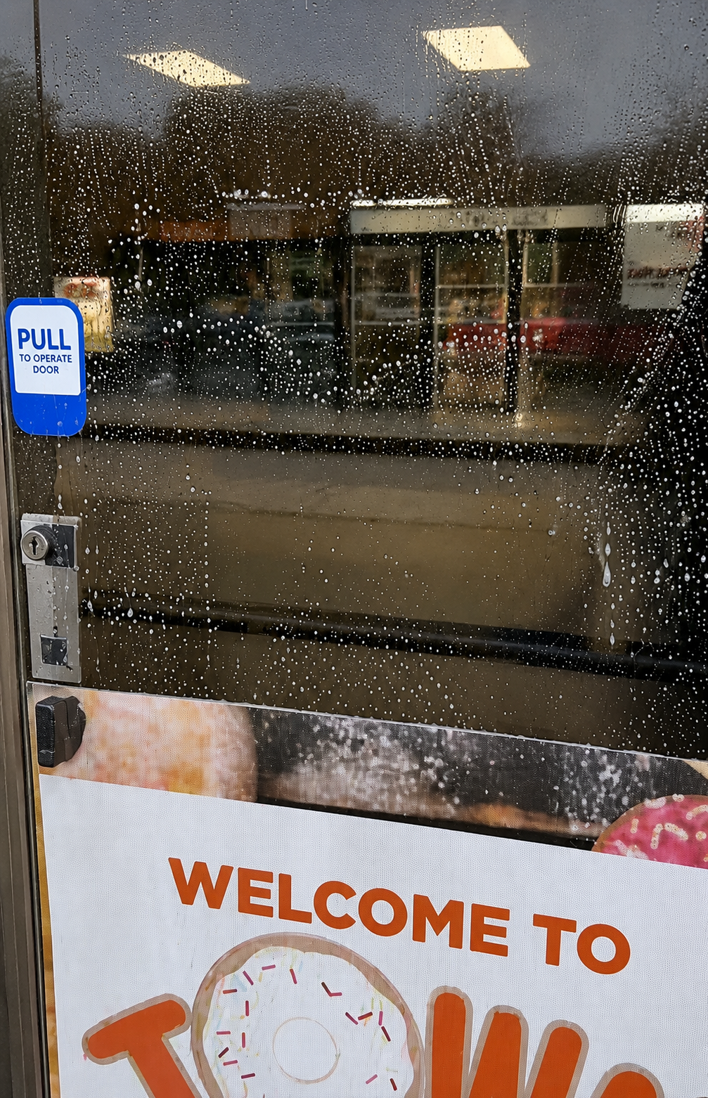 Rain-speckled glass door with a blue “PULL” sign and a “WELCOME TO” poster inside a store.