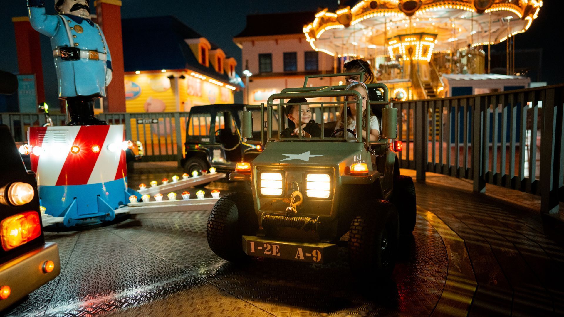 Military jeep ride at a carnival. Lights, carousel, and buildings in the background.