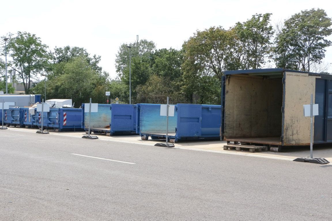 Blue and beige dumpsters lined up outdoors on asphalt, with trees in the background.