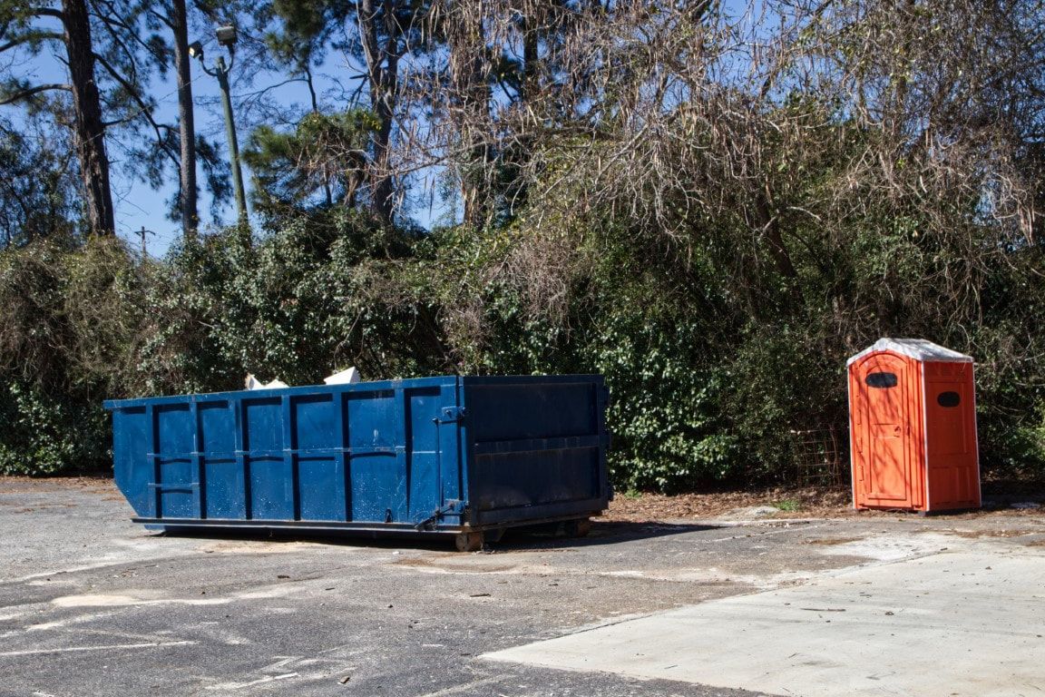 Blue dumpster next to an orange portable toilet, both on a paved area with trees in the background.