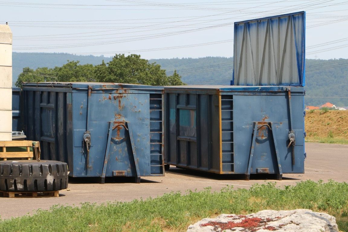 Blue metal dumpsters, one with an open lid, on a paved area with a tire and grassy foreground, hills in the background.