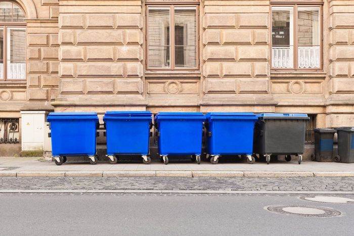 Blue and dark gray trash bins line a sidewalk in front of a tan building with windows.