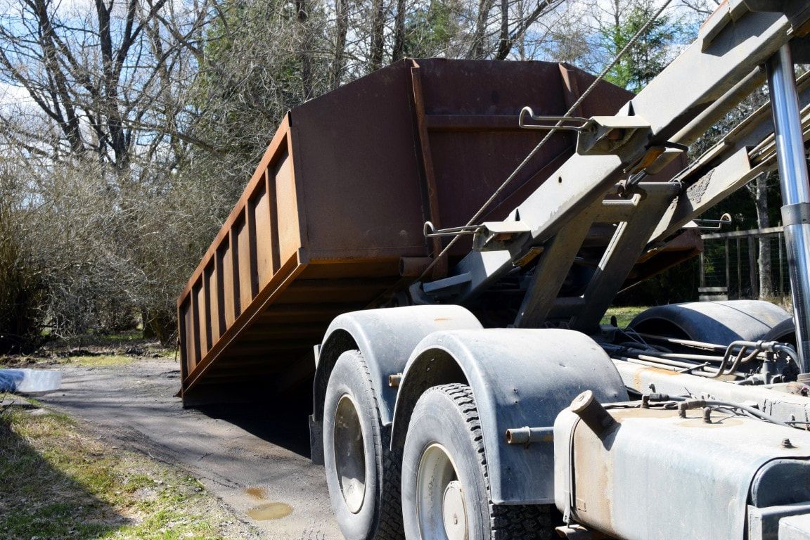 Brown dumpster being lifted by a truck in a driveway. Sunny day, trees in background.