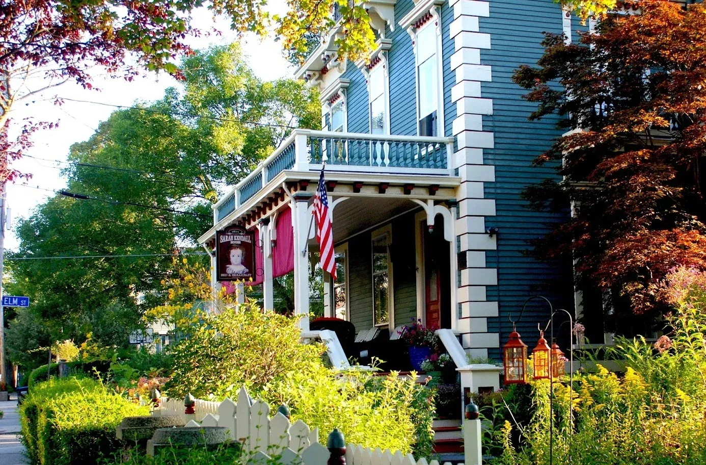 Victorian house with blue siding, white trim, and a porch surrounded by lush garden greenery.