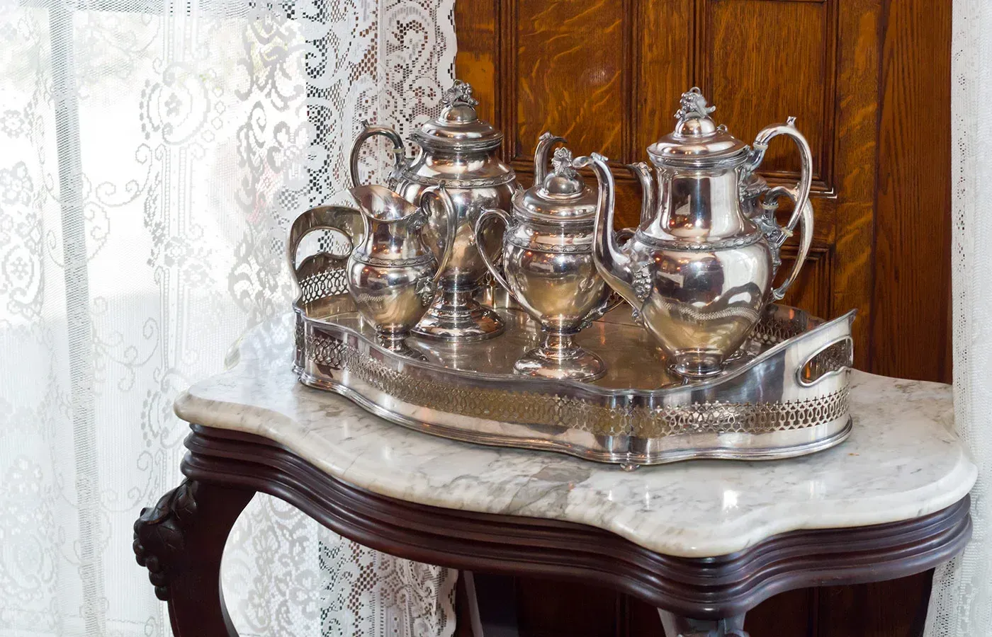 Silver tea service on a tray atop a marble-topped wooden side table near lace curtains