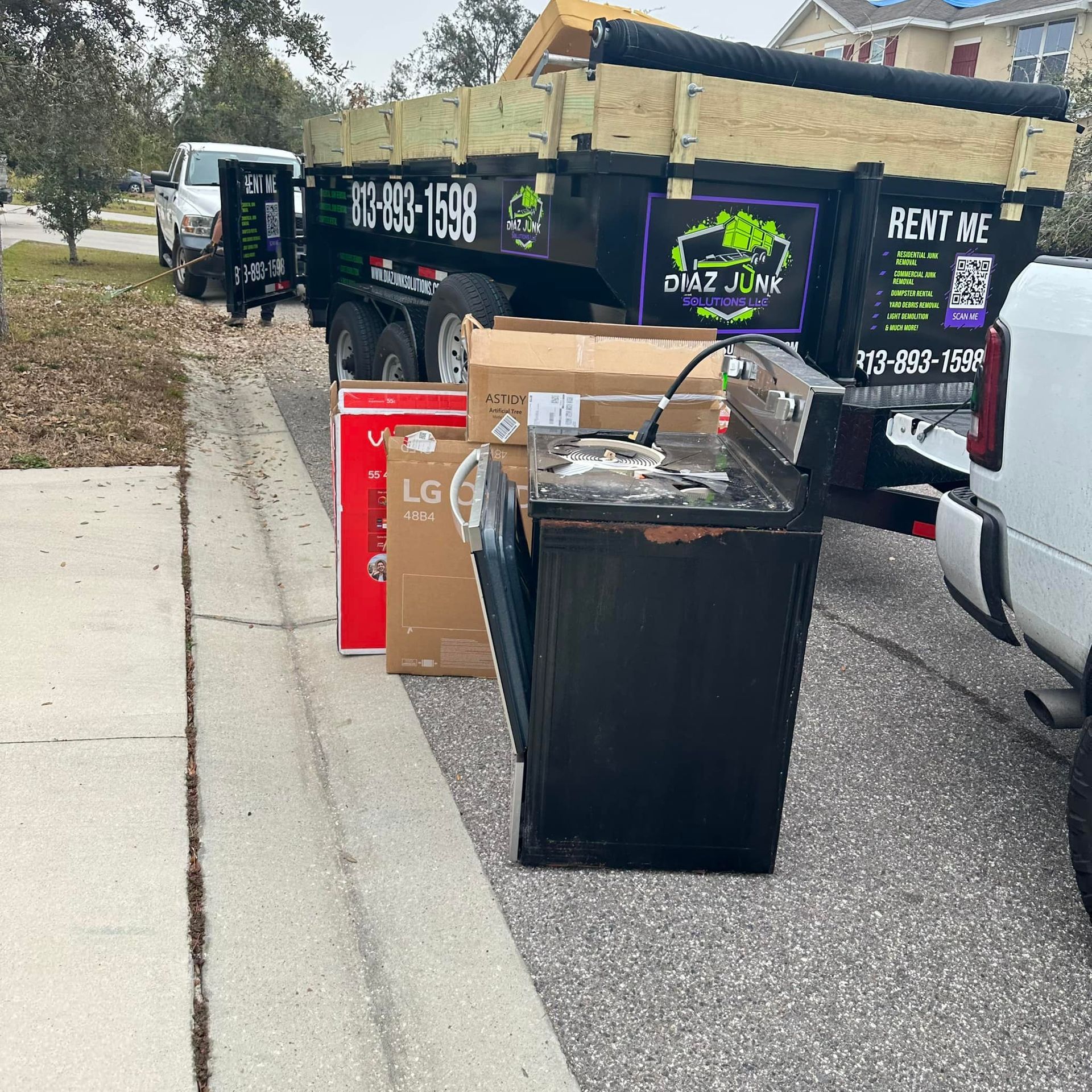 A black dumpster trailer and two pickup trucks loaded with trash and appliances, on a residential street.