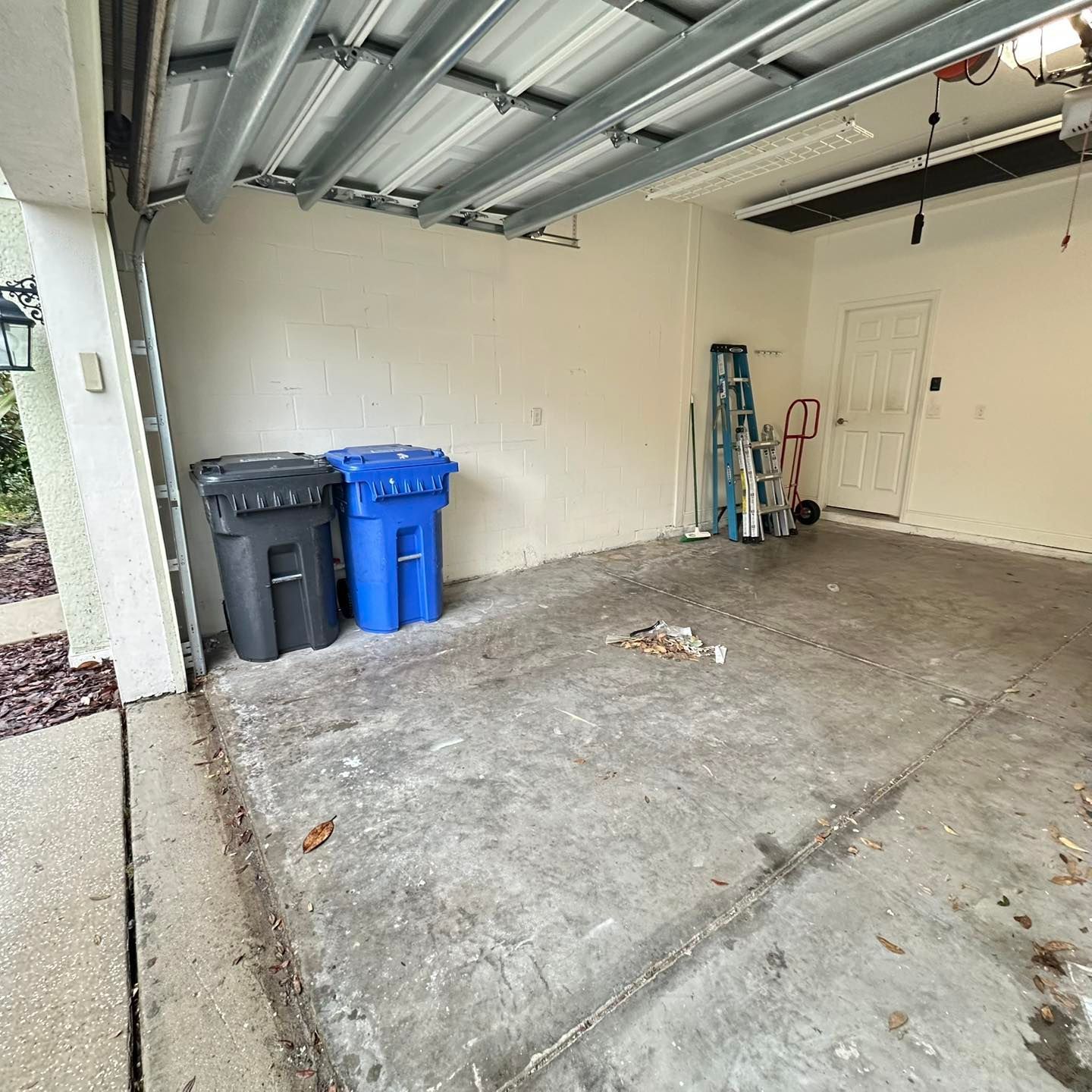 Garage with trash cans, ladder, and white door. Open garage door, concrete floor, beige walls.