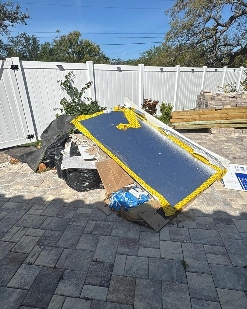 Pile of construction debris in backyard. Includes large reflective sheet, cardboard, and trash bag, near a white fence.