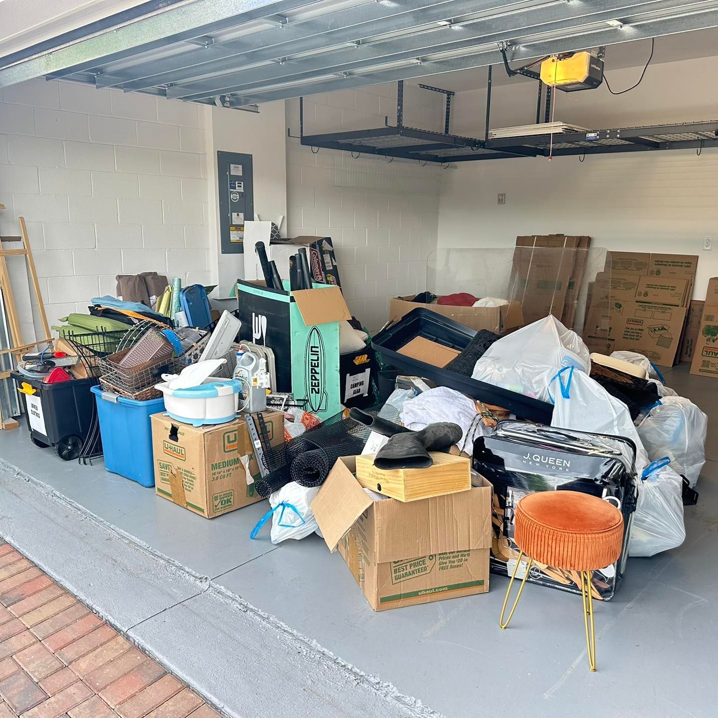 Cluttered garage with boxes, bags, and items stacked on the floor. An orange stool sits on the concrete surface.