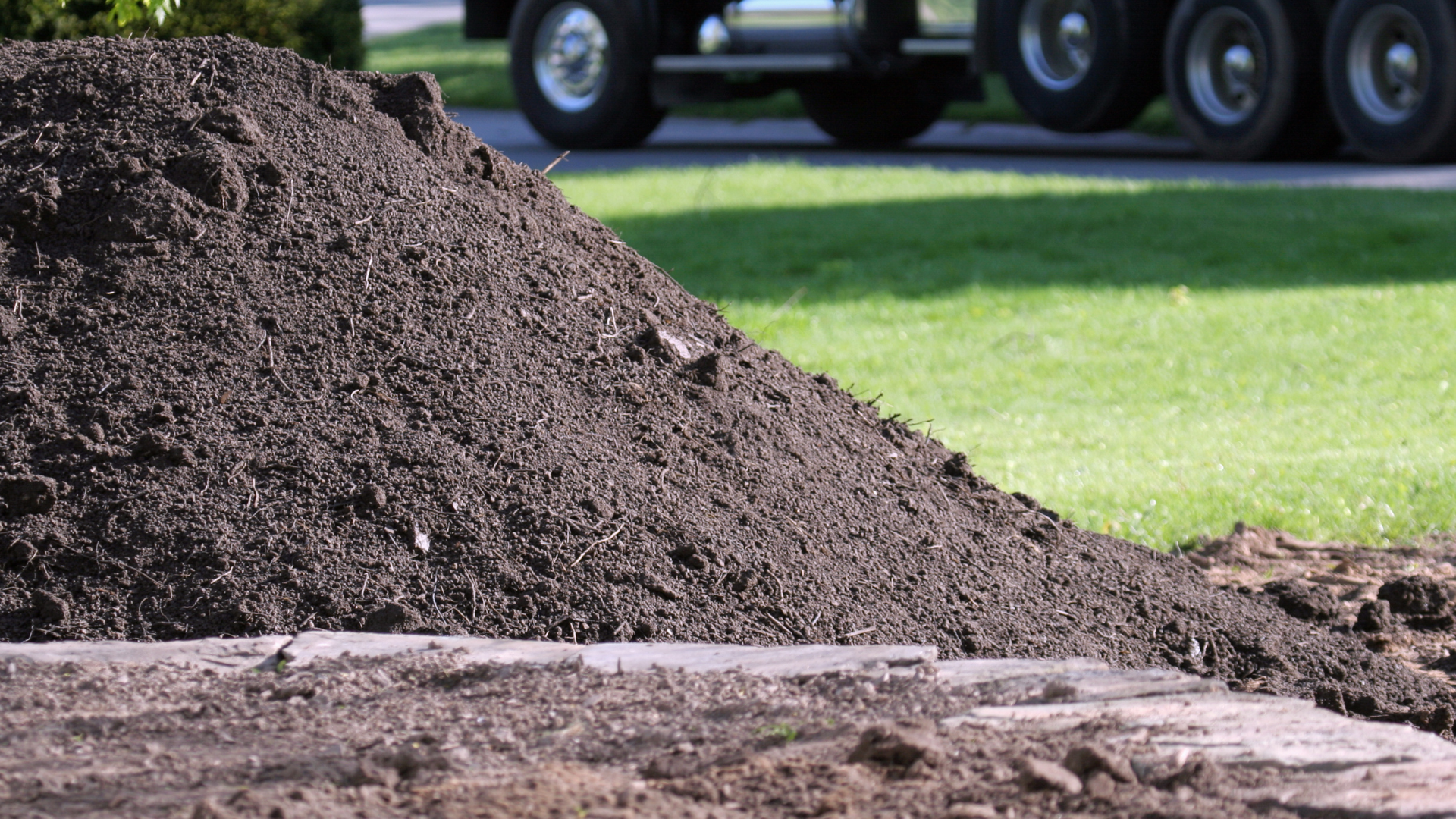 Pile of dark soil on the edge of a lawn, with truck wheels visible in the background.