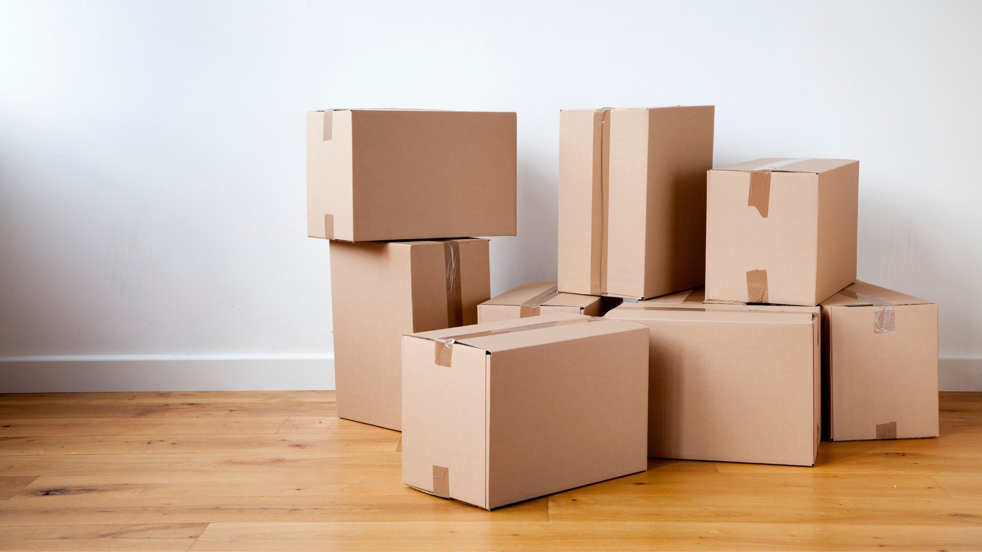 Cardboard moving boxes stacked on wooden floor against a white wall.