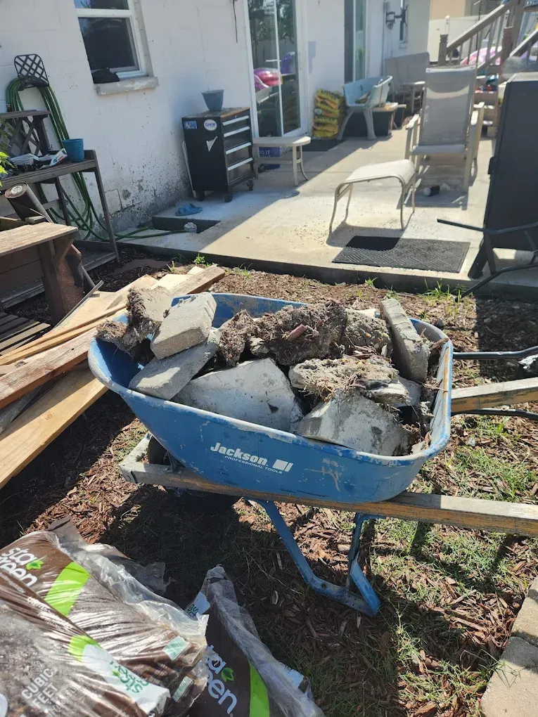 Blue wheelbarrow filled with rocks and debris, sitting in a backyard. A bag of mulch is in front.