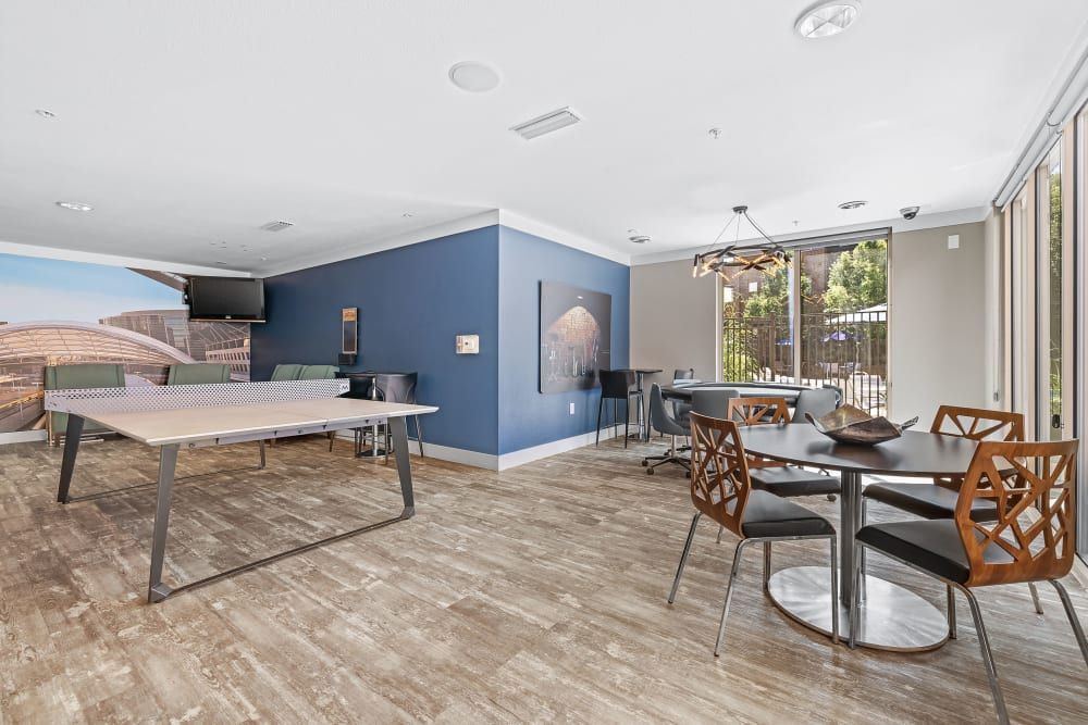 Modern multifunctional room with recreational and dining areas featuring a ping-pong table, bar-height seating by a kitchenette, a dining table with stylish chairs, and large windows offering natural light at Marq Inverness in Englewood, CO.