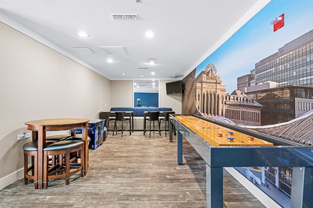 Modern game room with a wooden round table and stools on the left, a large shuffleboard table in the center, and high bar stools against a kitchen counter in the background. The room features a spacious feel with wood-look flooring, recessed lighting, and a large wall mural of city buildings on the right-hand wall, contributing to an urban atmosphere at Marq Inverness in Englewood, CO.