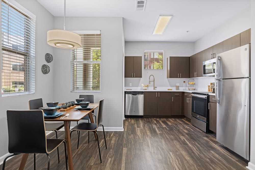 Modern kitchen and dining area with natural light. Features include stainless steel appliances, wooden cabinetry with a neutral color palette, a dining table set for four with dark chairs, and coordinated modern dishware. Decorative elements include a hanging cream light fixture over the dining table and circular wall art. Large windows with blinds offer a view of the exterior landscape at Marq Inverness in Englewood, CO.
