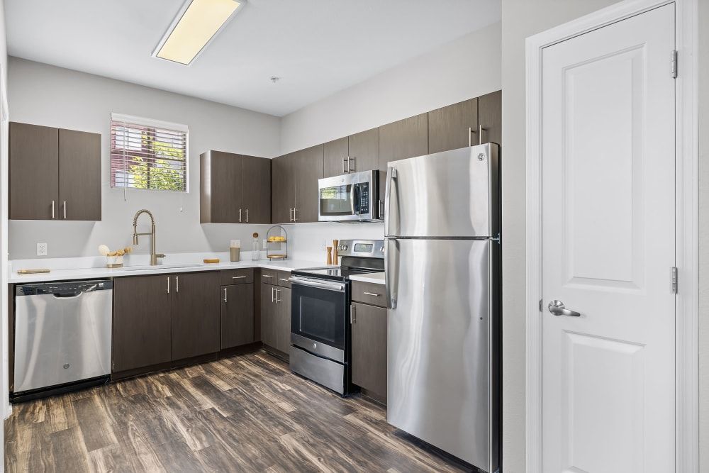 A modern kitchen with dark wood cabinets, white countertops, and stainless steel appliances, including a dishwasher, stove, and fridge. A window with blinds lets in natural light, and the floor is covered with dark wood-like laminate. A white door is on the right side of the frame at Marq Inverness in Englewood, CO.