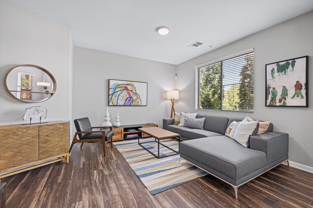 Modern living room interior with a gray sectional sofa adorned with assorted throw pillows. A wooden floor is partially covered by a striped area rug, and a wooden coffee table is centered in the room. To the left, a decorative gold-patterned sideboard is below a round mirror, with abstract and figurative artworks hanging on the walls. Natural light pours in from a large window with blinds on the right side of the room at Marq Inverness in Englewood, CO.