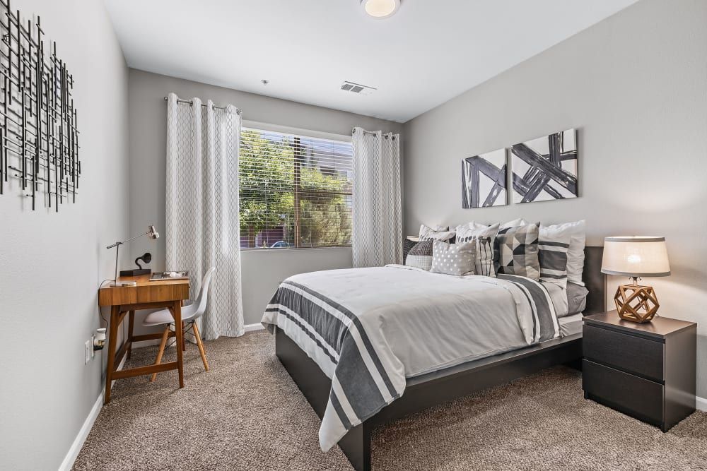 A well-lit modern bedroom featuring a neatly made bed with striped and patterned gray bedding. To the left, there is a small work desk with a wooden chair and a desk lamp, and on the wall hangs a geometric metal wall art. The right side showcases a nightstand with a lamp and a decorative piece, next to a triptych of abstract wall paintings. Large windows with white curtains bring natural light into the room with a view of greenery outside. The room has a plush carpet flooring and a neutral color palette at Marq Inverness in Englewood, CO.