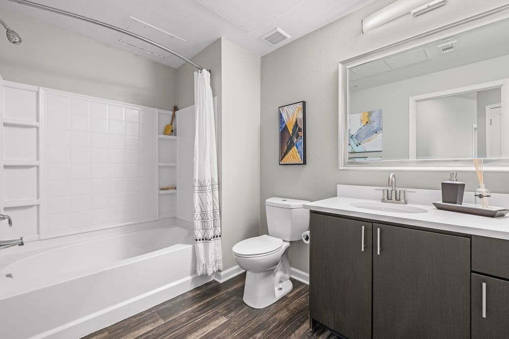 Modern bathroom interior with a white bathtub and shower curtain on the left, a toilet beside it, and a dark wood vanity with a white countertop and an integrated sink on the right. Above the vanity is a large mirror reflecting the opposite wall, and next to it hangs a colorful abstract painting. The room has wood-like flooring and is painted in light and neutral colors, creating a clean and bright atmosphere at Marq Inverness in Englewood, CO.