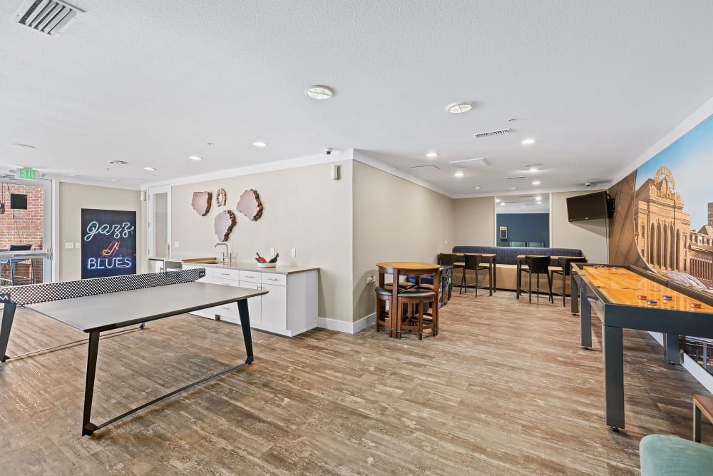 Modern recreational room with a ping pong table in the foreground, a shuffleboard table to the right, high bar stools by a counter, and a neon 'Jazz & Blues' sign on the wall. The room has wooden flooring and is well-lit with recessed ceiling lights at Marq Inverness in Englewood, CO.
